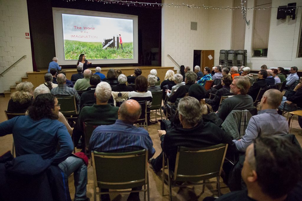 Vue de l'arrière d'une salle remplie de personnes assises qui regardent une conférencière sous une photo projetée sur un écran.