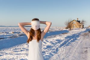 Une jeune fille vêtue d'un drap blanc enveloppe son visage dans un tissu blanche, elle se tient dans un décor hivernal, sur le bord du fleuve, une maison à l'arrière-plan.