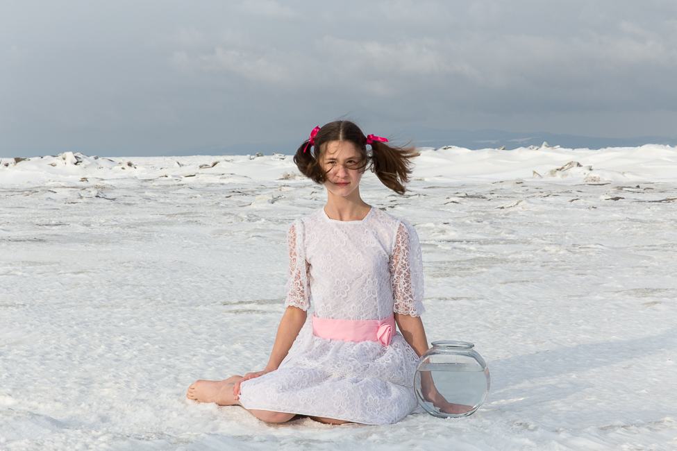 Une adolescente en robe d'été blanche est assise sur une banquise de neige au côté d'un bocal de poisson remplie d'eau, le vent balaie ses cheveux.