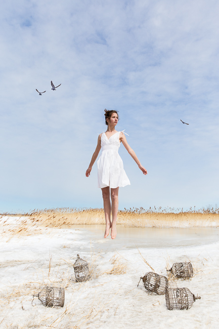 Teenage girl wearing a white summer dress in elevation above a snow covered field with empty bird cages beneath her.