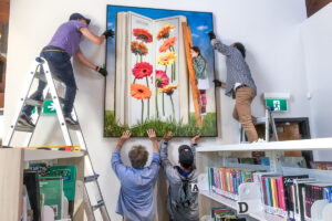 Men from behind install a huge photograph on a wall overhanging bookshelves.