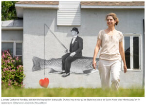 À l'avant plan une femme sourit en marchant dans l'herbe, derrière elle un photomontage d'un homme qui pêche sur une truite est affiché sur un mur de maison.