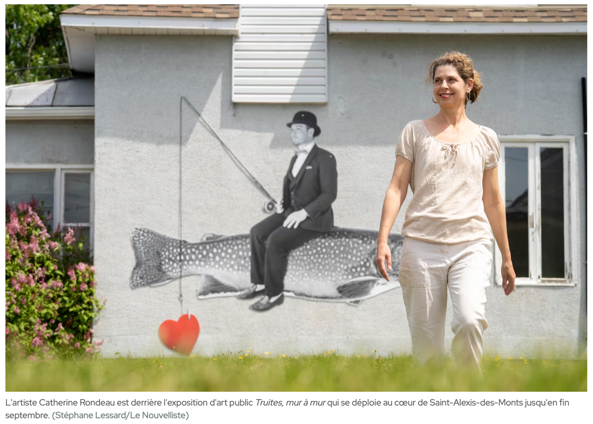 À l'avant plan une femme sourit en marchant dans l'herbe, derrière elle un photomontage d'un homme qui pêche sur une truite est affiché sur un mur de maison.