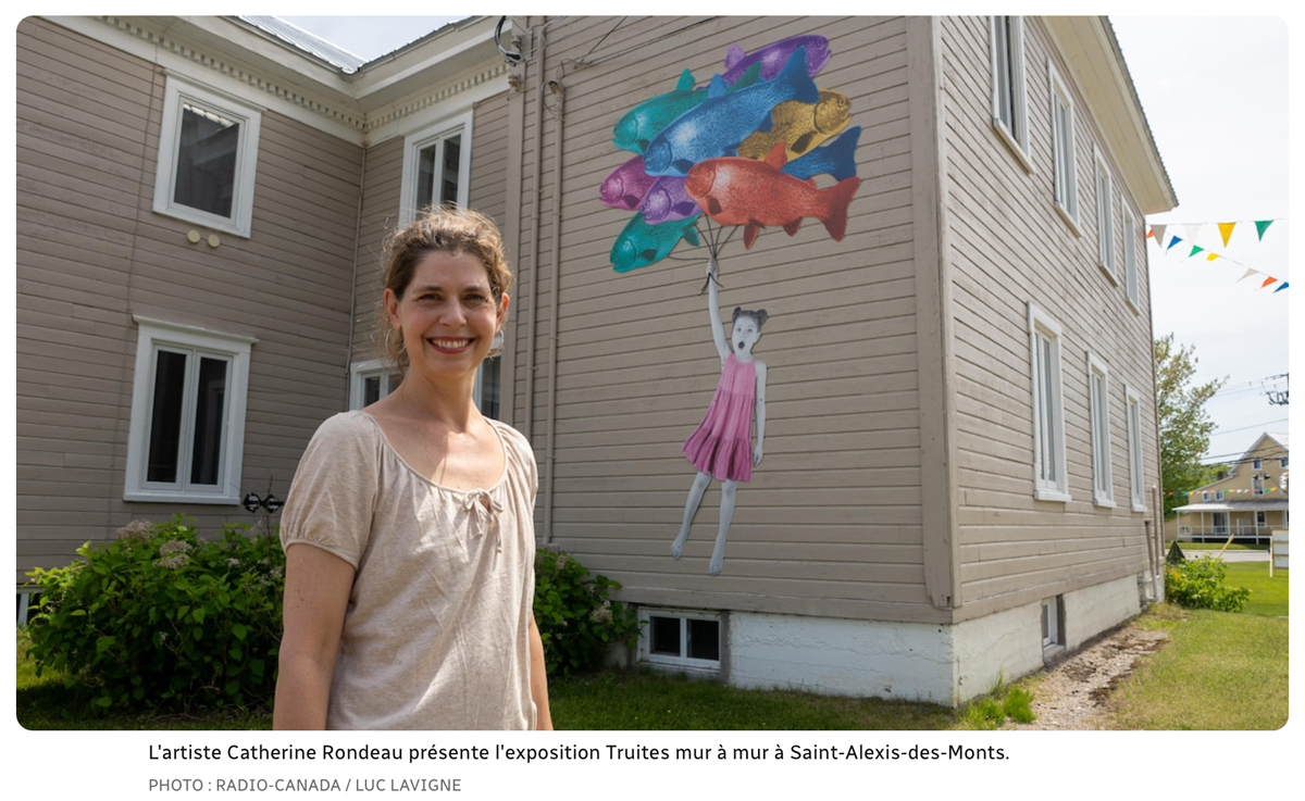 À l'avant-plan une femme sourit à la caméra. Derrière elle un photomontage d'une enfant qui s'envole en tenant un bouquet de ballons en forme de poissons est accroché au mur extérieur d'une maison.