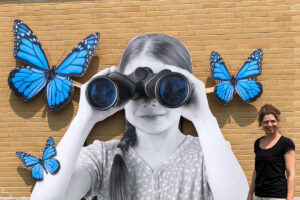 L'artiste Catherine Rondeau se tient devant une grande photographie en noir et blanc d'une enfant qui regarde dans des jumelles avec d'immenses papillons bleus qui volent autour d'elle.