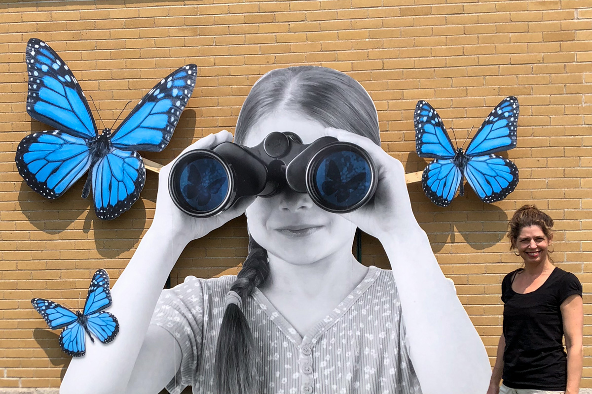L'artiste Catherine Rondeau se tient devant une grande photographie en noir et blanc d'une enfant qui regarde dans des jumelles avec d'immenses papillons bleus qui volent autour d'elle.