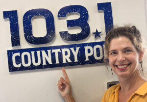 Une femme souriante pointe du doigt une enseigne qui se lit : 103,1 COUNTRY POP